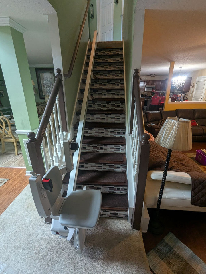 Bruno Elan stairlift at the foot of a carpeted staircase beside a living room lamp