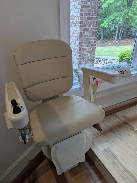 Handicare stairlift chair with a beige seat beside a window overlooking a garden