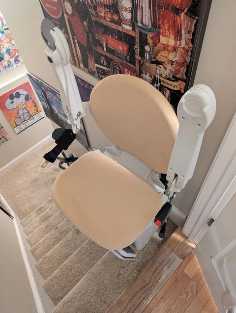Handicare stairlift with a beige seat at the top of carpeted stairs, framed art on the wall