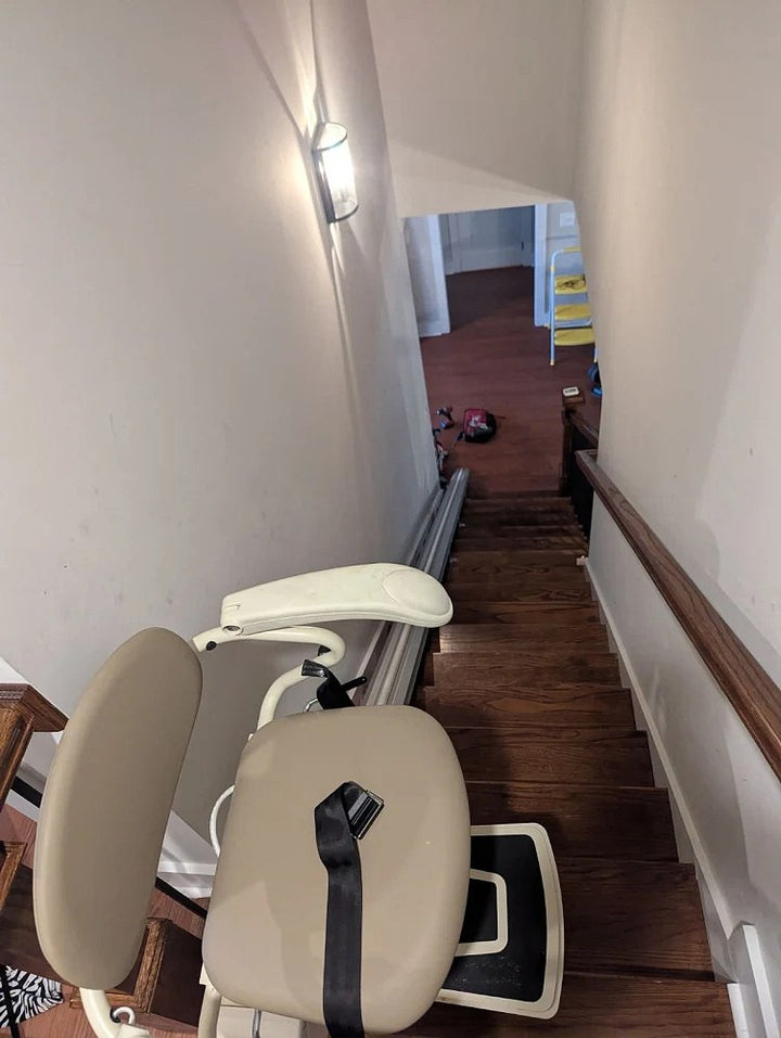 Harmar stairlift with a beige seat and seat belt at the top of narrow wooden stairs