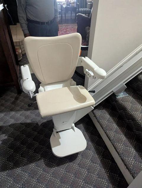 Stairlift with a cream seat at the foot of carpeted stairs in a living room