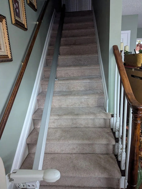 Stairlift rail along carpeted stairs next to a wooden banister, chair folded at the bottom