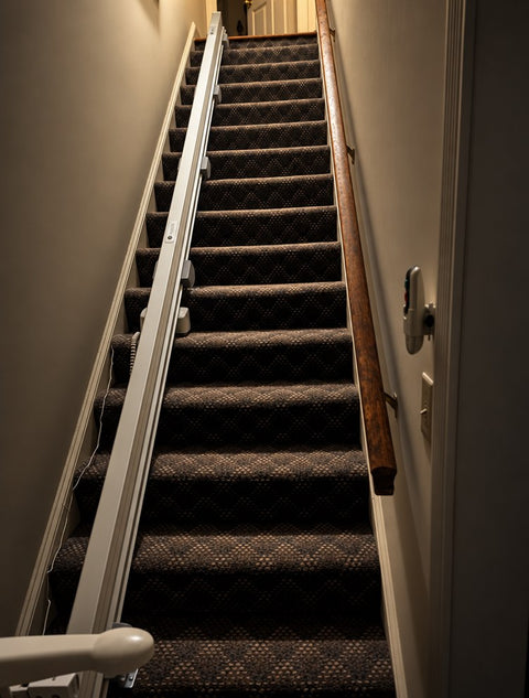 Stairlift rail along dark patterned carpeted stairs with a wooden handrail