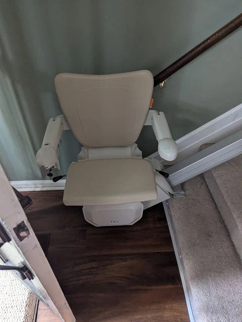 Stairlift with a beige seat parked at the top of stairs beside a wooden handrail