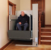 Man in a wheelchair on a Bruno residential platform lift (VPL-3100/3200B series shown) beside indoor stairs